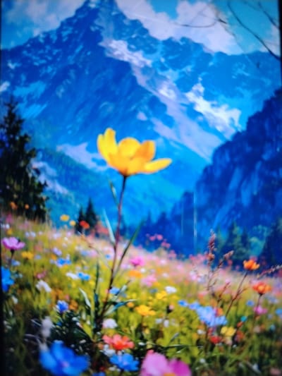 A vibrant, blurry image depicts a meadow filled with a multitude of colorful wildflowers in the foreground, set against a backdrop of majestic, blue, snow-capped mountains. A single yellow flower is prominently featured in the middle ground, its petals catching the light. The scene evokes a sense of natural beauty and tranquility. There is no indication of the city of Qorowul, Uzbekistan, within the image.