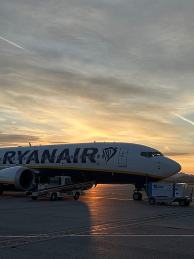 A Ryanair airplane is parked on the tarmac at Orio al Serio Airport, Italy. The plane is being serviced by a baggage truck and an Easy Load truck, with an orange sunset sky in the background.