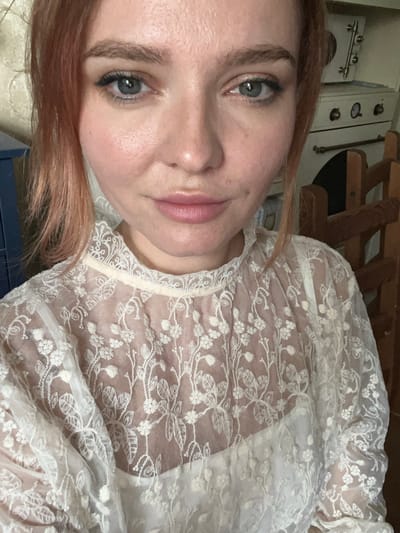 The media file is a close-up, low-angle portrait of a young Caucasian woman with light skin and reddish-brown hair. She is wearing a sheer white lace top with floral embroidery. Her left eye is visible, showing a light blue iris and dark eyelashes. Her lips are slightly parted, and she has a neutral expression. The background is out of focus, but appears to be an indoor setting with wooden furniture and what looks like a kitchen appliance in the upper left corner. There are no visible text elements in the image. The lighting suggests it is daytime.