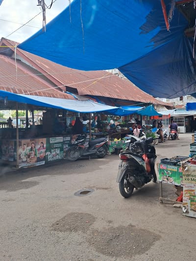 This image depicts an outdoor market scene in Dayahbaro, Indonesia, characterized by various stalls and vendors. The market is covered by large blue tarpaulins, providing shade. In the foreground, a dark-colored motorcycle is parked. Behind it, a stall features large advertisements with images of women and text in Indonesian, likely promoting food products. Several other motorcycles are visible throughout the market, suggesting transportation for vendors or customers.

Various stalls display goods, including fruits and vegetables, with some vendors and customers interacting. People are seen wearing traditional Indonesian attire, including headscarves. The ground is paved with asphalt and appears somewhat wet in places, with puddles visible. In the background, more market stalls and buildings are discernible, indicating a bustling commercial area. The overall environment suggests a typical Indonesian market, active with daily commerce.