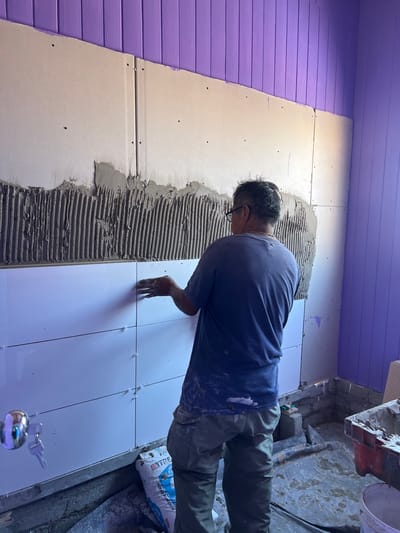 The image captures a scene of a person actively involved in tiling a wall, likely in an indoor construction or renovation setting. The perspective is from slightly above and behind the person, angled to the right.
The central figure is an adult, appearing to be a male, seen from his back and right side. He is wearing a dark blue short-sleeved t-shirt and olive green or khaki-colored long pants. His clothing shows smudges or splashes of white material, likely from the tile adhesive or grout. He has short, dark hair and is wearing glasses. He is leaning forward, focused intently on the wall he is working on, with his right hand positioned near a newly placed tile.
The wall itself is composed of different sections. To the left, there is a section of horizontally-oriented wooden planks or siding painted a vibrant lavender or light purple color. This section meets a lighter-colored substrate, possibly drywall or cement board, which forms the surface for the tiling. This substrate has several small screw or nail holes visible.
On the lighter wall section, a thick layer of gray tile adhesive has been applied in textured horizontal ridges, indicating it was spread with a notched trowel. To the right of this prepared section, several shiny white, rectangular tiles have already been installed. Small, white plastic tile spacers are visible between the tiles, ensuring uniform gaps for grout. The person's right hand is placed on or near one of these recently installed tiles, suggesting he is pressing it into place or adjusting its position.
The floor beneath the work area appears rough and unfinished, likely concrete, with some scattered debris indicative of a construction site. On the far right, near the bottom, a white bag with blue text is partially visible. The word "CIMENTO" (Portuguese for cement) is clearly readable on the bag, along with other indistinct text, suggesting it contains building materials like cement or tile adhesive. In the bottom foreground, slightly out of focus, there is a small, reflective metallic object, possibly a spherical bell, with keys attached.
The lighting in the scene appears bright, possibly from artificial sources, illuminating the work area. The overall impression is one of dedicated manual labor during a home improvement or construction project.