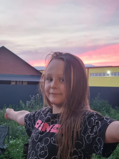 This is a vertical, close-up, outdoor photo of a young girl with long, brown hair. She is looking at the camera with a slight smile. She is wearing a black t-shirt with a pattern of white outlines of cartoon characters, possibly Mickey Mouse, and pink lettering that reads "PINK". The background shows a vibrant pink and orange sky, suggesting it is either sunrise or sunset. Behind her, there is a red-tiled roof of a building, a dark blue wall, and green foliage. A yellow building with a row of windows is visible in the upper right portion of the background. The girl is positioned in the foreground, with her head and upper body filling most of the frame. The lighting suggests it is dusk or dawn. The location is specified as Искра, Russia.