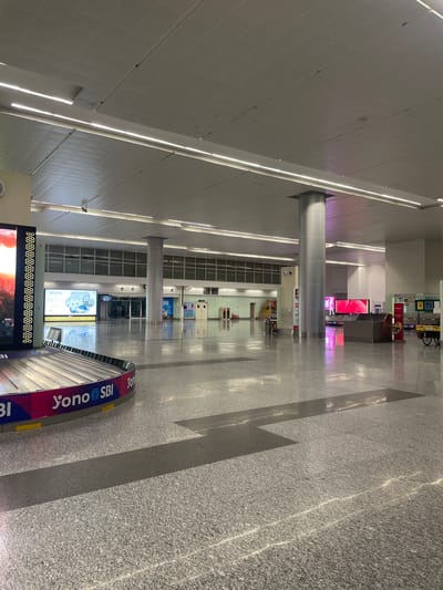 The image depicts the interior of a large, modern airport terminal, likely Raipur Khurd, India, given the presence of "Yono SBI" branding on the baggage claim carousel. The space is characterized by its expansive, polished granite floor, reflecting the bright, diffused lighting from long, linear fixtures embedded in the suspended ceiling. Several large, cylindrical grey pillars support the ceiling and divide the open space.

In the foreground, a baggage claim carousel is prominently visible on the left, featuring bold "Yono SBI" branding in pink and blue. To the left of the carousel, a large digital display screen shows a vibrant image of a cityscape at sunset.

The midground shows a wide, empty expanse of the terminal floor, leading towards a series of entrances and information displays. A large digital billboard on the left displays an advertisement for "RAN BAAS" with imagery of hotel rooms and amenities. Further back, the terminal appears to have a section with large windows and what looks like reception desks or service counters. There are a few people visible in the distance, appearing as small figures near the entrances, suggesting low passenger traffic at the time of the photograph.

The environment is clean and well-maintained, typical of an airport terminal. The lighting is bright and even, creating a sense of openness and spaciousness. The overall impression is one of a modern, functional transportation hub.