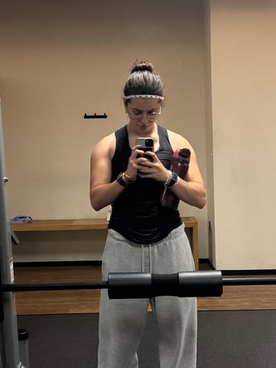 In Komchén, Mexico, an individual is captured in a mirror selfie within a gymnasium setting. The subject, a person with dark hair pulled into a bun and wearing a white patterned headband, round eyeglasses, a black sleeveless top, and light gray sweatpants, stands facing a mirror. They hold a dark-colored smartphone in both hands at chest level, indicating the act of taking the photograph. A dark red water bottle is tucked under their left arm. Accessories include multiple bracelets and a smartwatch on the left wrist, and a watch on the right wrist.
The background features a plain light beige wall. Three black wall-mounted hooks are visible on the left side of the wall, above a light brown wooden bench or shelf. A purple towel rests on the left end of this bench. The foreground is occupied by gym equipment: a vertical metal gym rack structure on the far left, and a black barbell with a central black padded squat pad positioned horizontally across the lower frame, in front of the subject. The floor is dark gray, consistent with gym flooring.