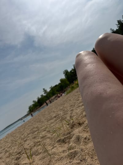 The image depicts a close-up, low-angle view of a person's legs resting on a sandy beach. The legs, which appear to be wet and glistening, dominate the foreground and right side of the frame. The beach extends into the background, with a body of water visible on the left, reflecting the sky. Scattered individuals can be seen along the shoreline and in the water, engaged in various beach activities. Dense green foliage borders the beach in the mid-ground, with trees forming a backdrop against a partly cloudy sky. The scene captures a typical summer day at a lakeside or seaside location. The image does not contain any specific visual cues that would allow for precise location identification within Aiyrtau, Kazakhstan.
