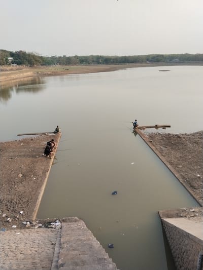 This is an eye-level, medium shot photograph taken at a reservoir in Blimbing, Indonesia. The reservoir is the dominant feature, with calm, murky water occupying most of the frame. The sky is overcast and pale.

In the foreground, there is a concrete structure extending into the water. On this structure, a group of four individuals are seated, fishing. They are positioned on the left side of the concrete platform. To the right, another concrete structure also extends into the water, with a single individual fishing on it. This individual is seated on a wooden plank. The concrete structures appear weathered and have dirt and some debris scattered on them.

In the background, the far bank of the reservoir is visible, lined with trees and vegetation. Further in the distance, there are some buildings. The overall impression is of a quiet, somewhat neglected reservoir. The water itself contains some scattered debris.