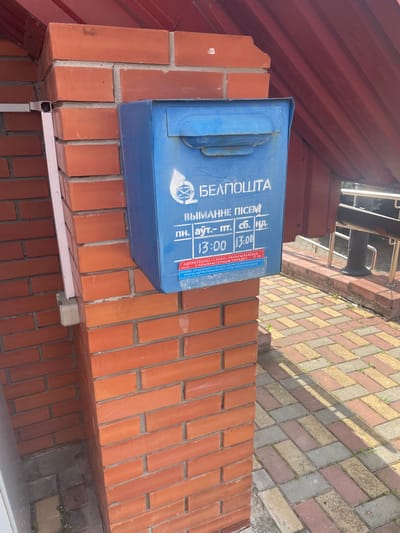 A blue mailbox is mounted on a red brick pillar. The mailbox has text in white lettering, "БЕЛПОШТА," and an emblem featuring a winged envelope. Below this, it lists operating hours: "ВЫМАННЕ ПІСЕМ" (Attention Mail), followed by "пн. — пт. сб. нд." (Mon. — Fri. Sat. Sun.) and time slots "13:00 13:00". A smaller red sticker at the bottom reads "АПРАЦУЮЦЦА Ў" (Processed) followed by more text that is difficult to decipher. The pillar is adjacent to a paved walkway with a repeating pattern of rectangular pavers in shades of red, yellow, and gray. A dark red, corrugated roof structure is visible above and to the right of the mailbox. The environment appears to be outdoors, possibly in a residential or public area.