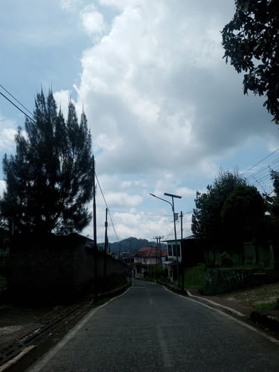 The image depicts a street in Ban Phouviengnoy, Laos. The road is asphalt, with curbing on both sides. Buildings line the street, and trees are present on both sides. Overhead power lines are visible. The sky is cloudy, with patches of blue visible. A mountain can be seen in the distance. There are no people visible in the image.