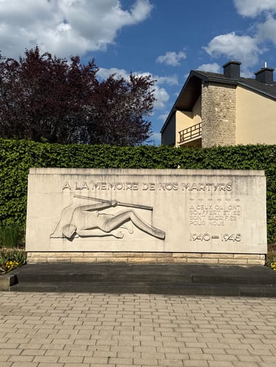 This outdoor shot features a stone monument in Manternach, Luxembourg. The monument, dedicated "A LA MEMOIRE DE NOS MARTYRS" (To the Memory of Our Martyrs), has a relief carving of a fallen figure. Text on the monument further indicates it honors those who suffered and sacrificed themselves for the nation between 1940 and 1945. The monument is framed by hedges and greenery, and a corner of a building is visible on the left, along with a tree. The foreground includes a stone-paved area. The sky is partially cloudy.