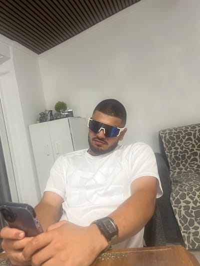 In an indoor setting, a man wearing a white t-shirt and large white-framed sunglasses is seated at a wooden table. He is holding a smartphone in his left hand, with his right hand resting on the table. A dark-colored wristwatch is visible on his right wrist. Behind him, a patterned sofa is visible. To his left, a white cabinet with two doors is present, topped with decorative items including figurines and a small plant. The ceiling features a striped design. The location is identified as Rosenovo, Bulgaria.