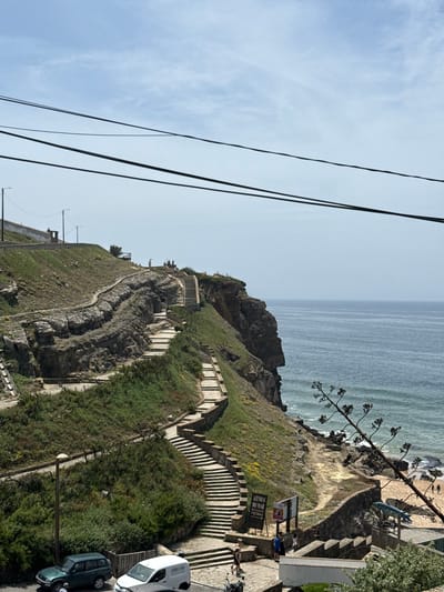 This image depicts a scenic coastal cliffside with a winding stone staircase leading down towards a beach and the ocean. The staircase is integrated into the rocky terrain, with green vegetation and scattered yellow wildflowers growing on the slopes. At the top of the cliff, there are paved pathways and a few people can be seen enjoying the view. The ocean is a calm, blue expanse under a partly cloudy sky. In the foreground, several vehicles, including a dark green SUV and a white van, are parked on a paved area. A small sign with Portuguese text, possibly indicating directions or local attractions, is visible near the base of the stairs. The scene appears to be located in the Azenhas do Mar area of Colares, Portugal, a location known for its dramatic cliffside houses and ocean vistas.