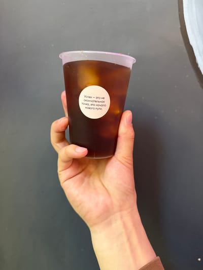 A person's hand holds a clear plastic cup filled with a dark liquid and ice. A circular white sticker with text in Russian is affixed to the cup. The background is a dark, textured surface. The location of this scene in Veshki, Russia, is not discernible from the image.