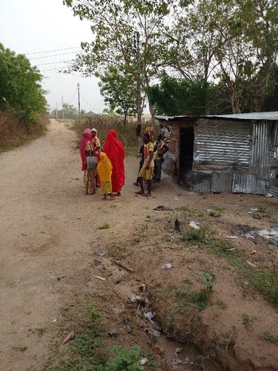 A group of people stand on a dirt road in a rural setting. Three women wear brightly colored robes, while two men stand nearby, clad in casual clothing. A small corrugated metal structure sits to the right, likely a shelter or storage space. Trash litters the ground in a nearby ditch. The scene evokes a sense of poverty and simplicity, juxtaposed with the vibrant colors of the women's attire. Overhead are utility lines, hinting at some infrastructure in the area.