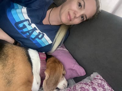 A person lies on a gray surface, likely a sofa or bed, with a dog lying on a purple pillow beside them. The person has fair skin and blonde hair, wearing a blue GAP brand shirt. The dog is brown, white, and black with a pink collar. Another patterned pillow is adjacent to the purple pillow. The setting appears to be indoors. No location details indicate Danilov, Russia.