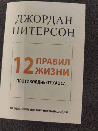 The image is a close-up, slightly angled shot of the cover of a book. The book cover is predominantly white with text in dark gray and orange. At the top, in large, serif font, is the name "ДЖОРДАН ПИТЕРСОН" (Jordan Peterson). Below this, a box outlines the number "12" in a large, orange font, followed by "ПРАВИЛ" (Rules) and "ЖИЗНИ" (of Life) in smaller orange text. Underneath this, in dark gray text, it reads "ПРОТИВОЯДИЕ ОТ ХАОСА" (Antidote to Chaos). At the bottom of the cover, in smaller dark gray text, is "ПРЕДИСЛОВИЕ ДОКТОРА НОРМАНА ДОЙДЖА" (Foreword by Dr. Norman Doidge). The background of the image is a textured, dark gray surface, likely fabric. There is no indication of the city of Schwäbisch Gmünd, Germany, or any environmental context beyond the book cover and the surface it is resting on.