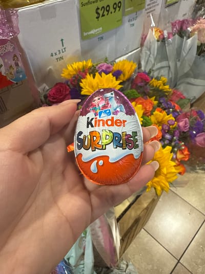 A hand holds a Kinder Surprise egg against a backdrop of a bouquet of flowers and what appears to be a cardboard box with a green price tag indicating "$29.99 Sunflower Oil Brookhaven 4x3LT TIN". The location, based on the "Brookhaven" label, is likely a grocery store in Brookhaven, Darien, United States of America. The image does not show specific people or actions beyond the holding of the Kinder Surprise egg. The environment suggests a purchase or gifting scenario, with the flowers and chocolate treat as possible presents.