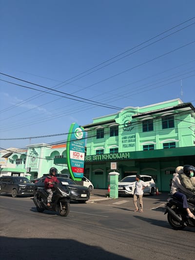 The image depicts the RS SITI KHODIJAH hospital in Pekalongan, Indonesia, on a clear day. The hospital building is painted a bright green and features multiple windows and architectural details. In the foreground, a black motorcycle with a rider wearing a maroon jacket and a helmet is driving on the road. To the right, another motorcycle with two passengers is also on the road. A white SUV is parked on the side of the road near the hospital entrance, and a man in a light-colored shirt and pants is standing on the sidewalk. Traffic lights and street signs are visible, including a large green sign for the hospital's IGD (Emergency Department). Power lines are strung across the sky above the scene.