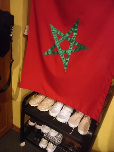 A partially visible Moroccan flag drapes over a shoe rack filled with an assortment of sneakers and athletic shoes. The flag features a prominent red background with a green, five-pointed star. Beneath the flag, a black purse or bag with a shoulder strap rests on a wooden surface. The shoe rack is dark and has a multi-tiered design, accommodating several pairs of shoes. The background consists of a yellow-toned wall and wooden flooring.