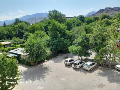 In the city of Kara-Köl, Kyrgyzstan, under a clear blue sky with distant mountains, this image captures a paved outdoor area. The scene features a gravelly parking lot with several cars: a white SUV, a white sedan, and another white SUV. Adjacent to the parking area is a small, single-story building with a metal roof and green trim, possibly a garage or storage unit, partially covered by dense green trees. The surrounding environment is lush with numerous trees and vegetation, suggesting a rural or semi-rural setting within the city. Shadows from the trees are cast across the ground, indicating the time of day is likely late morning or early afternoon, as suggested by the timestamp "10:50" in the lower left corner. The timestamp also indicates the date as "June 29, 2026". The overall impression is one of a peaceful, sun-drenched day in a verdant landscape.