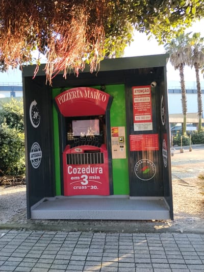 This image depicts a self-service pizza vending machine, branded "PIZZERIA MARCO," situated outdoors in Viana do Castelo, Portugal. The kiosk has a dark exterior frame with bright green interior panels and a prominent red central section containing the machine's interface. Above, tree foliage with hanging roots casts shadows, while in the background, palm trees stand under a clear, sunny sky, indicating daytime. The ground in front of the kiosk is paved with grey interlocking concrete blocks.
The machine advertises "24h" service and "ARTESANALE" (artisanal) quality. A large red sign on the machine indicates "Cozedura em 3 min" (Cooking in 3 min), suggesting pizzas are cooked fresh upon order. Below this, "cruas ™ 30." likely refers to raw pizzas being available or dispensed in 30 seconds. On the right panel, a red warning sign, "ATENÇÃO" (ATTENTION), states that "A MÁQUINA FUNCIONA EM PRÉ-RESERVA" (The machine works on pre-reservation). It clarifies that it "SÓ COBRA SE A PIZZA FOR ENTREGUE EM CASO DE PROBLEMA NÃO COBRA NADA" (It only charges if the pizza is delivered. In case of a problem, it does not charge anything). An emergency contact number, "922 102 444," is also listed. A smaller, red sign below the warning sign likely provides a menu or additional instructions.
The machine features a central screen, slots for payment (coins/cards), and a dispensing opening. A round logo with "PIZZERIA MARCO" and a pizza slice graphic is visible on the lower right side of the kiosk. No people are present, highlighting the automated vending process.