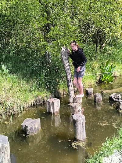 A young girl stands barefoot on a wooden post in a shallow stream, part of an obstacle course at a recreational area in Sint Maartensvlotbrug, Netherlands. She is wearing a black hoodie and gray shorts, and holding onto a taller wooden post for balance. The stream is filled with several other wooden posts, creating a path across the water. Green grass and trees line the banks of the stream.