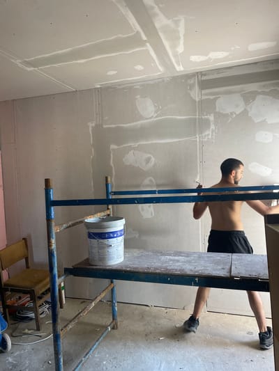 In a room with unfinished drywall, a young man, bare-chested and wearing black shorts and athletic shoes, is working. He is positioned behind a blue metal scaffolding with a wooden plank surface. On the plank sits a large white bucket with blue lettering, likely containing joint compound or paint. The man is holding a tool, possibly a trowel or putty knife, and appears to be applying material to the wall behind him. The walls and ceiling are covered in drywall panels, with visible seams and patching compounds. To the left, a wooden chair with a worn seat is visible, along with coiled electrical wires on the floor. The overall scene suggests a construction or renovation project in progress. The location is identified as Krushevo, Bulgaria.