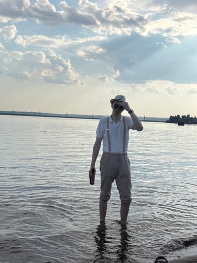 The photograph shows a man standing in shallow water in Пальники, Russia. The man is wearing a white shirt, tan trousers held up by suspenders, and a hat. He is also wearing sunglasses and a watch on his left wrist. He is holding a small, dark object in his right hand. His left hand is raised to his hat. The water is calm and reflects the sky. The sky is mostly blue with white clouds. There are trees in the distance.