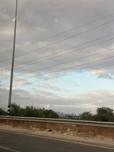 The image captures a view from a vehicle moving through the Sdot Dan Regional Council, Israel. The foreground is dominated by a paved road with a yellow line marking its edge. Adjacent to the road is a sandy embankment topped with a metal guardrail. Beyond the guardrail, there is sparse vegetation and scattered litter. In the middle ground, a dense line of green trees runs horizontally. In the background, a distant cityscape with numerous buildings is visible under a cloudy sky. A tall utility pole with black and white markings stands prominently on the left side of the frame, with several power lines stretching horizontally across the sky. The sky itself is a mix of gray clouds and patches of lighter blue. The overall impression is of a roadside scene on a partly overcast day, with a city skyline on the horizon.