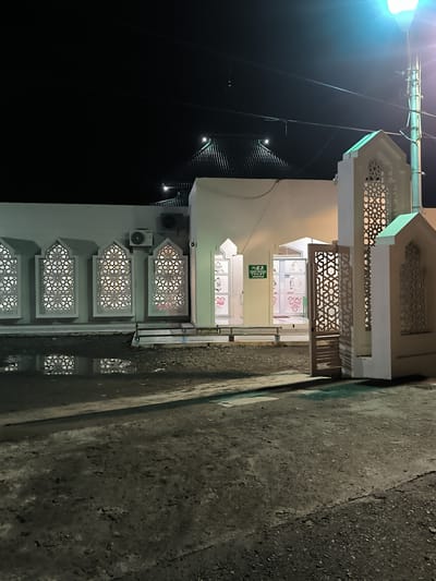 The image depicts the exterior of a building at night in Tibang, Indonesia. The building has white walls and features several decorative windows with intricate geometric patterns. An air conditioning unit is visible on the wall. In front of the building, there is a puddle of water reflecting the patterned windows. To the right, a decorative gate is open, revealing an entrance to the building. A sign with the word "TOILET" in green is also visible. The ground in front of the building appears to be unpaved and wet, with some gravel. A street lamp with a bright light is positioned to the right of the frame, illuminating the scene. The sky is dark, indicating nighttime.