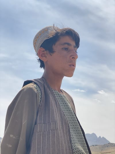 A medium shot depicts a young man, presumably of Afghan descent, dressed in traditional clothing. He wears a crocheted cap, a patterned shirt, and a pinstriped vest over a long-sleeved brown outer garment. His gaze is directed off-camera towards his right. In the blurred background, there are mountain peaks and a cloudy sky. The subject is photographed in Ashoqah, Afghanistan.