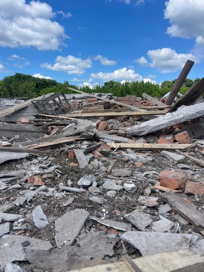 The image shows the debris field of a destroyed building. The debris is mostly concrete and wood. There are pieces of wood and metal, broken bricks, and small amounts of dirt and vegetation. The debris is scattered across a flat surface. The sky is blue with white clouds. This location is near the coordinates 50.979, 33.946, which is in the city of Terny, Ukraine. No people are visible in the image.