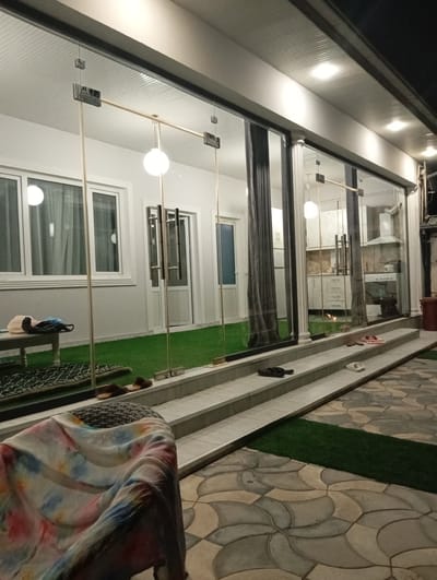 A nighttime exterior shot captures a modern building with large glass walls, revealing an interior that appears to be a kitchen and living area with artificial turf flooring. The building is lit from within and by external spotlights, creating a warm and inviting ambiance.

In the foreground, a patterned tiled patio is visible, with a narrow strip of artificial turf running alongside it. Several pairs of sandals and flip-flops are scattered on the steps leading up to the glass-enclosed area. A woven stool is partially visible, draped with a colorful, tie-dye-like fabric.

Through the glass, the interior is illuminated by soft lighting. The kitchen area features white cabinets, a stove, and a range hood. A dining area or living space is also visible, with white curtains and white doors. A large, round white light fixture hangs from the ceiling in the interior. The overall impression is one of a contemporary residential or commercial space, possibly a cafe or a home, designed for evening use. The time of day is clearly night, and the weather is not discernible from the image. The location is identified as Sasbaga, Uzbekistan.