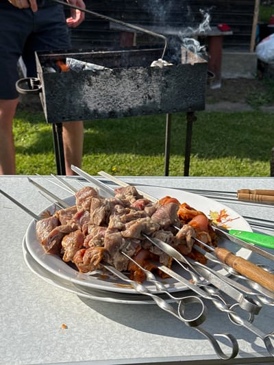 The image depicts a barbecue setting. On the left, a person is tending to a charcoal grill, with smoke rising from the burning fuel. The grill appears well-used, with soot and ash accumulated on its surface. On the right, a stack of white plates laden with skewers of meat is visible. The skewers are made of metal, and the meat consists of different cuts and seasonings. The setting is outside, and the city and country are unspecified.