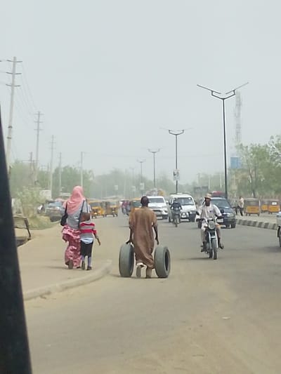 The image portrays a bustling street scene in Daurawa, Nigeria, under a hazy, light-gray sky. A paved multi-lane road with a central concrete median dominates the view. In the immediate foreground, a partial dark panel from a vehicle's interior is visible on the left. On the left side of the road, a female subject wearing a pink hijab and a multi-patterned dress walks away from the viewer, accompanied by a young child in a striped shirt and dark shorts walking beside her. Further into the road, a male subject in a brown robe (agbada) is observed from behind, manually pushing two large, dark vehicle tires along the asphalt, holding one in each hand. His feet are visible between the tires as he propels them forward. To the right of the individual with the tires, a motorcyclist, wearing a light-colored shirt and a light cap, operates a light-colored motorcycle, traveling towards the viewer. The road is populated by several other vehicles, including a white SUV, a dark sedan, a white van, and multiple yellow motorized rickshaws (tuk-tuks) positioned on both the left and right sides of the thoroughfare. The background features utility poles with overhead power lines on the left, a series of modern streetlights with distinctive V-shaped luminaires lining the road, and a tall communication tower visible on the far right. Sparse foliage and indistinct building structures are present in the distant background.