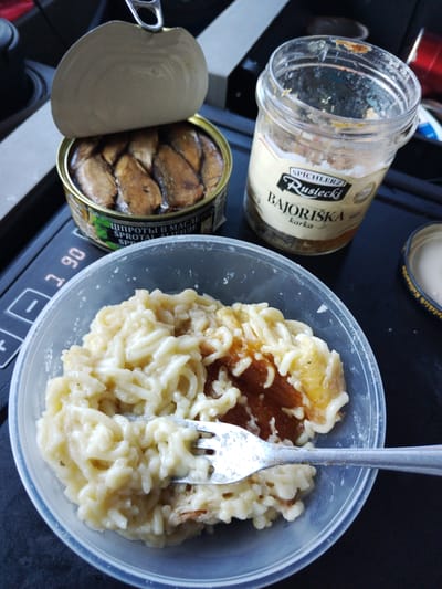 The image shows a close-up of a meal consisting of pasta with sauce and what appears to be some form of preserved fish. The meal is served in a clear plastic container. A fork is partially submerged in the pasta. To the left of the container is an open tin of sprats in oil, with several fish visible inside. To the right of the container is a partially empty glass jar with a label that reads "SPICHLERZ Rustecki BAJORISKA karka". The objects are placed on a dark surface that appears to be a tray or a table. A digital display showing "1.90" is visible on the left side of the image, possibly a price or a measurement. The scene does not provide any specific indicators of location within the city of Pforzheim, Germany, and therefore, this information cannot be included.