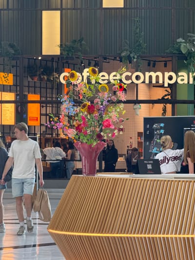 The image shows an indoor scene, likely a mall, in Leidschendam, Netherlands. In the foreground, a large, vibrant bouquet of artificial flowers is displayed in a deep red vase on a slatted wooden counter. The counter has an illuminated band of light beneath it. To the left, a young man wearing a white t-shirt and light blue shorts walks past, carrying two brown paper shopping bags. Behind him, a person's legs and lower torso are visible, also walking. In the midground, several people are visible interacting at a service counter or cafe. A large black digital screen displaying a mall map is visible behind them, with the text "MALL GUIDE" below it, and the Westfield logo. The background features a sign that reads "Coffeecompany" in large, illuminated letters, with hanging plants and shelving units visible behind it, suggesting a retail or food establishment. The overall lighting is a mix of ambient interior light and the illumination from the signs and the counter.