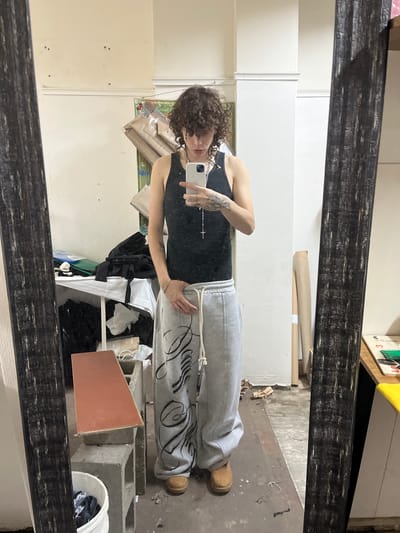 A person with curly hair is reflected in a dark-framed mirror, wearing a black tank top and gray sweatpants with a dark graphic design. The person is holding a white smartphone, presumably taking a selfie. The background behind the person in the reflection reveals a cluttered room. Several objects can be seen, including stacks of papers, a white shelf laden with black clothing, a terracotta tile propped against several gray cinder blocks, and a white bucket containing dark items. The flooring appears to be concrete. The location appears to be a disorganized domestic or storage space. The image does not contain sufficient information to determine its specific location in the city of Bahía Blanca, Argentina.