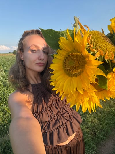 A young woman with light brown hair is lying in a field of grass with sunflowers. She is wearing a brown, ruched dress. The sunflowers are large and yellow, with some appearing wilted. The sky is clear and blue. The image appears to have been taken in the late afternoon or early evening, as indicated by the warm, golden light. The setting is a natural, outdoor environment, likely a field or meadow. The woman's face is visible and she is looking towards the camera with a neutral expression. The overall mood of the image is peaceful and serene.
