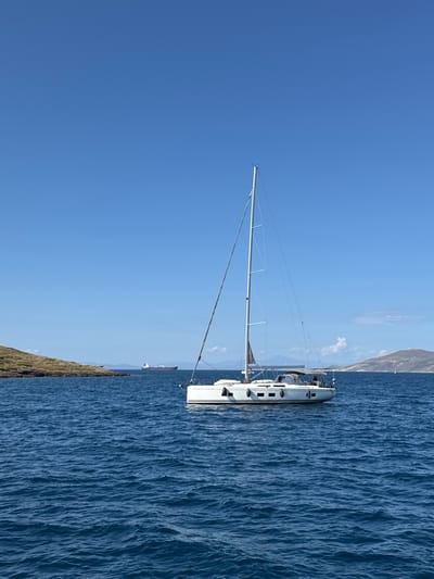 The image captures a maritime scene in the Lavreotiki Municipal Unit, Greece, featuring a white monohull sailboat on deep blue, choppy water under a clear, bright blue sky. The sailboat, positioned slightly to the right of the central axis, displays its mast in an upright position with sails furled or reefed. Several white fenders are visible along the port side of the vessel. A grey bimini or cockpit cover extends over the aft section, and at least one individual is discernable within the cockpit area.

To the left, a low-lying, rocky landmass with sparse, dry, brown-green vegetation is visible in the mid-ground. In the far distance, between this landmass and the distant horizon, a large dark-hulled cargo ship or tanker with red and orange elements is present on the water. A larger, hazy, mountainous or hilly landmass occupies the right side of the distant horizon, with additional faint landforms visible along the horizon line. The sky exhibits very few, scattered white clouds, primarily near the right horizon. The scene is illuminated by bright daylight, suggesting a midday or early afternoon setting.