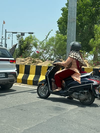 A woman wearing a pink outfit, red pants, and a patterned scarf is riding a black scooter on a sunny day in Ajmer, India. She is wearing a black helmet. The scooter is on a paved road, next to a white SUV. There is a yellow and black barrier in the background, with dirt and bushes behind it. A traffic light pole and trees are also visible.