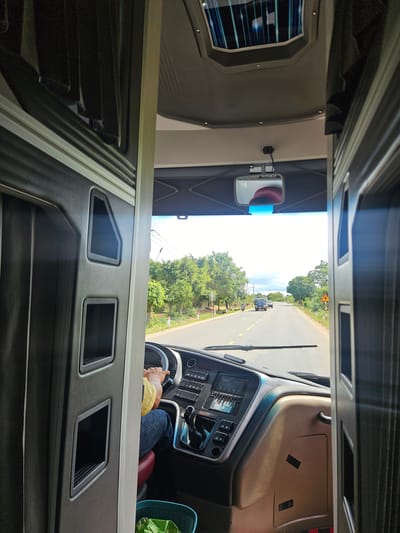 The image shows the interior of a bus, looking forward through the driver's cabin. A person wearing a yellow shirt and blue jeans is driving. The dashboard features a stereo system, various buttons, and a gear shift. Outside the bus, a paved road stretches ahead, lined with trees and vegetation. A truck and a motorcycle are visible on the road. The sky is bright and overcast. A small mirror is mounted on the ceiling, reflecting a portion of the driver's head. The bus appears to be traveling through a rural area, likely in Ia Khươl, Vietnam, based on the road and vegetation. The overall impression is that of a typical bus journey in a tropical region.