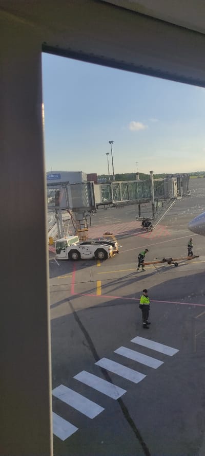 The image depicts an airport tarmac at Khrabrovo Airport in Kaliningrad, Russia, under a clear blue sky with scattered clouds. In the foreground, a white passenger boarding bridge connects to a building with "CARGO" signage. A white tow tractor, numbered "211," is parked on the tarmac. Several airport personnel in high-visibility vests are present. One individual is operating a piece of ground equipment, another is standing near it, and a third individual is further away. A pedestrian crossing is marked with white stripes on the tarmac. In the background, tall airport lighting poles and a distant treeline are visible. Part of an aircraft fuselage is also visible on the right side of the image.