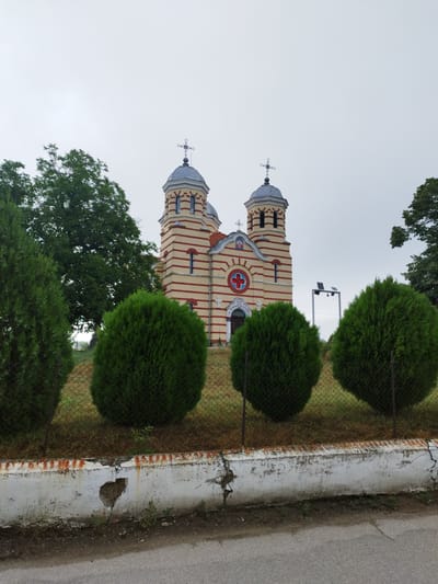 The image depicts a church in Slanotran, Bulgaria, under an overcast sky. The church is painted in alternating horizontal stripes of yellow and red. It features two bell towers, each topped with a dome and a cross. A large tree stands to the left of the church, and several round, green bushes are positioned in front of it, separated by a wire fence. A low, white concrete wall with a damaged section is visible in the foreground, with a road to its right.