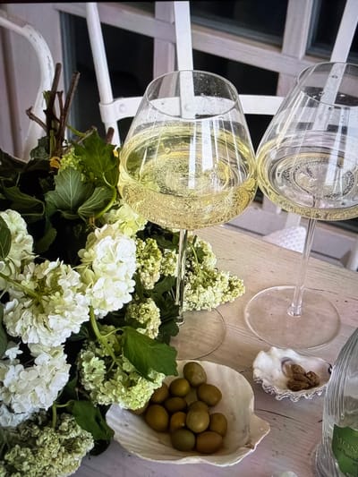 This close-up image, likely taken in Ros Na Rí, Ireland, captures an elegant outdoor or semi-outdoor setting arranged for a celebration or relaxed gathering. The primary focus is on two champagne flutes, both filled with a sparkling, light yellow beverage, suggesting champagne or prosecco. The glasses show condensation and numerous bubbles, indicating a chilled drink.

In the foreground and midground, a lush bouquet of flowers dominates the lower portion of the frame. The bouquet features clusters of white and light green blossoms, possibly hydrangeas or viburnum, intertwined with dark green leaves, adding a fresh and natural touch to the scene.

To the right, resting on a light-colored wooden or whitewashed table surface, are two decorative shells. One is a fluted white shell containing what appears to be a small cluster of walnuts or similarly textured small brown items. Below it, another larger, smooth white shell holds a generous serving of green olives. Partially visible behind these items is a clear glass bottle with a green label, on which the text "MON" can be discerned, suggesting part of a brand name. Another empty, clear wine glass stem is also visible on the table.

The background features white, slatted furniture or architectural elements, possibly part of a patio railing or chairs, creating a bright and airy atmosphere. The natural light suggests it is daytime. The overall arrangement conveys a sense of sophistication, relaxation, and anticipation of an enjoyable event.