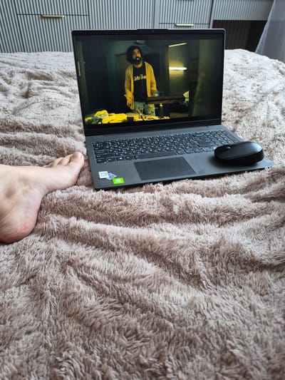 A person's bare foot is visible on the left side of the image, resting on a fluffy, beige carpet. To the right of the foot, a closed laptop is positioned on the same carpet. The laptop screen displays a still image of a man with dark, curly hair and a beard, wearing a yellow cardigan over a dark shirt and blue jeans. He appears to be standing indoors, with a dimly lit background. A black computer mouse is placed on the right side of the laptop. The background behind the carpet shows a striped wall, possibly part of a room's decor. The overall setting appears to be a casual indoor environment, likely a bedroom or living room. The image was captured in Bilohorodka, Ukraine.
