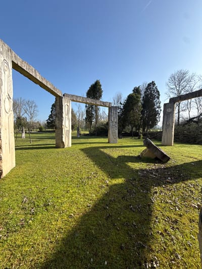 This image depicts an outdoor sculptural park located in Mealhada, Portugal, under a clear blue sky. The scene is dominated by several large, light-colored, rough-textured concrete or stone structures resembling primitive-style gateways or archways. Each structure comprises two vertical pillars supporting a horizontal lintel. The pillars feature carved abstract, anthropomorphic, or symbolic figures and patterns.
In the foreground, a large, rectangular concrete block lies tilted on the grass, supported by a smaller stone. Moving further into the scene, an arrangement of three complete gateway structures is visible, with additional pillars or fragments scattered in the background. The ground is covered by vibrant green grass, interspersed with scattered dry leaves. Tall, slender evergreen conifer trees, along with bare deciduous trees, populate the background and midground. Strong sunlight casts prominent, elongated shadows of the structures across the grassy terrain. A single, faint white contrail is visible in the upper center-left portion of the clear blue sky. No individuals are present in the scene.