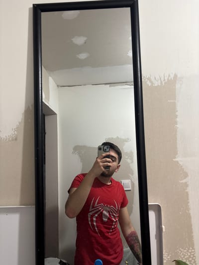 An interior scene in Erden, Bulgaria, depicts a male subject capturing a mirror selfie. The individual, possessing short dark hair and light facial stubble, wears a red short-sleeved t-shirt with a prominent white Spider-Man logo. He holds a light-colored smartphone in his right hand, obscuring the lower portion of his face, to photograph his reflection in a tall, black-framed rectangular mirror. A multicolored tattoo, appearing as a floral or tree-like design, is visible on his left forearm. The background, both reflected in the mirror and partially visible outside its frame, consists of walls in an apparent state of renovation or unfinished decoration, characterized by patches of lighter plasterwork, uneven paint application, and a white electrical switch or outlet. The reflected ceiling also shows visible repairs. A partially obscured white rectangular object is visible in the lower right reflection. The environment appears utilitarian and under construction.