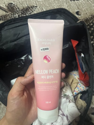 This media file shows a close-up of a hand holding a pink tube of body cleanser. The tube is from the brand KWAILNARA and is labeled "MELLOW PEACH BODY CLEANSER" in English and Korean. The text on the tube also includes hashtags like "#달콤피치" (sweet peach), "#촉촉보습" (moisturizing), and "#산뜻거품" (refreshing foam). Below this, there is a description in English: "The peach body cleanser gives a pleasant feeling with its Soft and Moisturizing. Its exotic scent makes your body enjoy and lovely." The tube also indicates it contains "180 ml" and has a price of "₩2,000".
The background of the image shows the inside of a black bag, with some colorful patterned fabric and other items visible. The lighting suggests it might be indoors or under artificial light. The hand holding the tube appears to be that of an adult. No specific time of day, weather, or strong emotional cues are evident, beyond the product itself suggesting a focus on self-care and pleasant sensations. The location context provided is Tashlak, Kyrgyzstan, which is not directly discernible from the image itself.