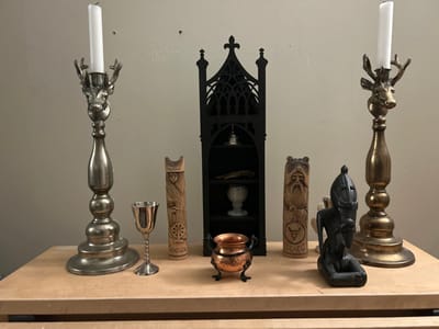 The image displays a static arrangement of ritualistic or decorative objects on a light-colored wooden surface against a plain, light-colored wall. Two small, dark circular marks are visible on the wall above the center of the arrangement. The scene is located in the city of Gränsta, Sweden.
From left to right, the objects are:
1. A tall, silver-toned metal candlestick, featuring a stag's head and antlers, holding an unlit white taper candle.
2. A small, polished silver-colored metal goblet with a decorative stem.
3. A rectangular, light-toned wooden carving depicting a stylized female figure holding a spoked wheel, with partially visible carved text beneath.
4. A small, black, gothic-style open cabinet with three shelves. The top shelf contains a small silver-colored bell; the middle shelf holds an elongated, irregular brown object; the bottom shelf displays a light-colored cup.
5. A small, copper-toned cauldron with a single handle, resting on a black three-legged stand, positioned in front of the black cabinet.
6. A rectangular, light-toned wooden carving depicting a bearded male figure with bear-like features, a symbol resembling a bull's head with a triangle, and partially visible carved text beneath.
7. A dark-colored, possibly black, figurine depicting a kneeling or squatting human-like figure with an elongated headpiece.
8. A tall, brass-toned metal candlestick, featuring a stag's head and antlers, holding an unlit white taper candle.
No people or actions are depicted in the image.