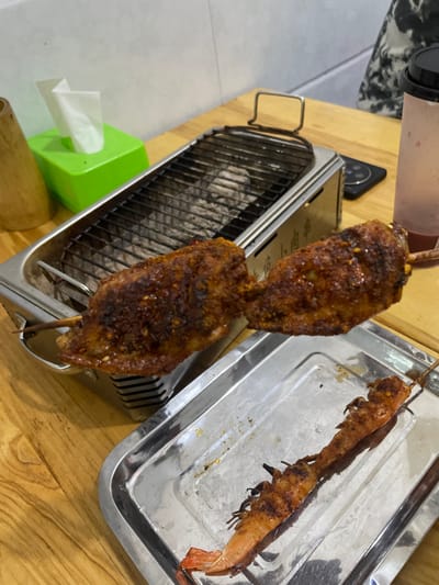 The scene depicts a tabletop dining area within an establishment in Dali City, China. Dominating the view are two grilled chicken wings on a wooden skewer, heavily crusted with red spices, positioned centrally over a rectangular stainless steel tabletop charcoal grill. The grill's grate covers visible charcoal embers and ash. Below, on a reflective rectangular metal tray, lies a single grilled prawn skewer, exhibiting orange-red coloration and char marks. Environmental elements include a green tissue dispenser holding white tissues and a tall, cylindrical bamboo container positioned on the left side of the wooden table. On the right, a plastic cup with a dark lid and straw holds a reddish liquid, alongside a dark-colored smartphone. A partial view of an individual's patterned attire is discernible in the upper right background.